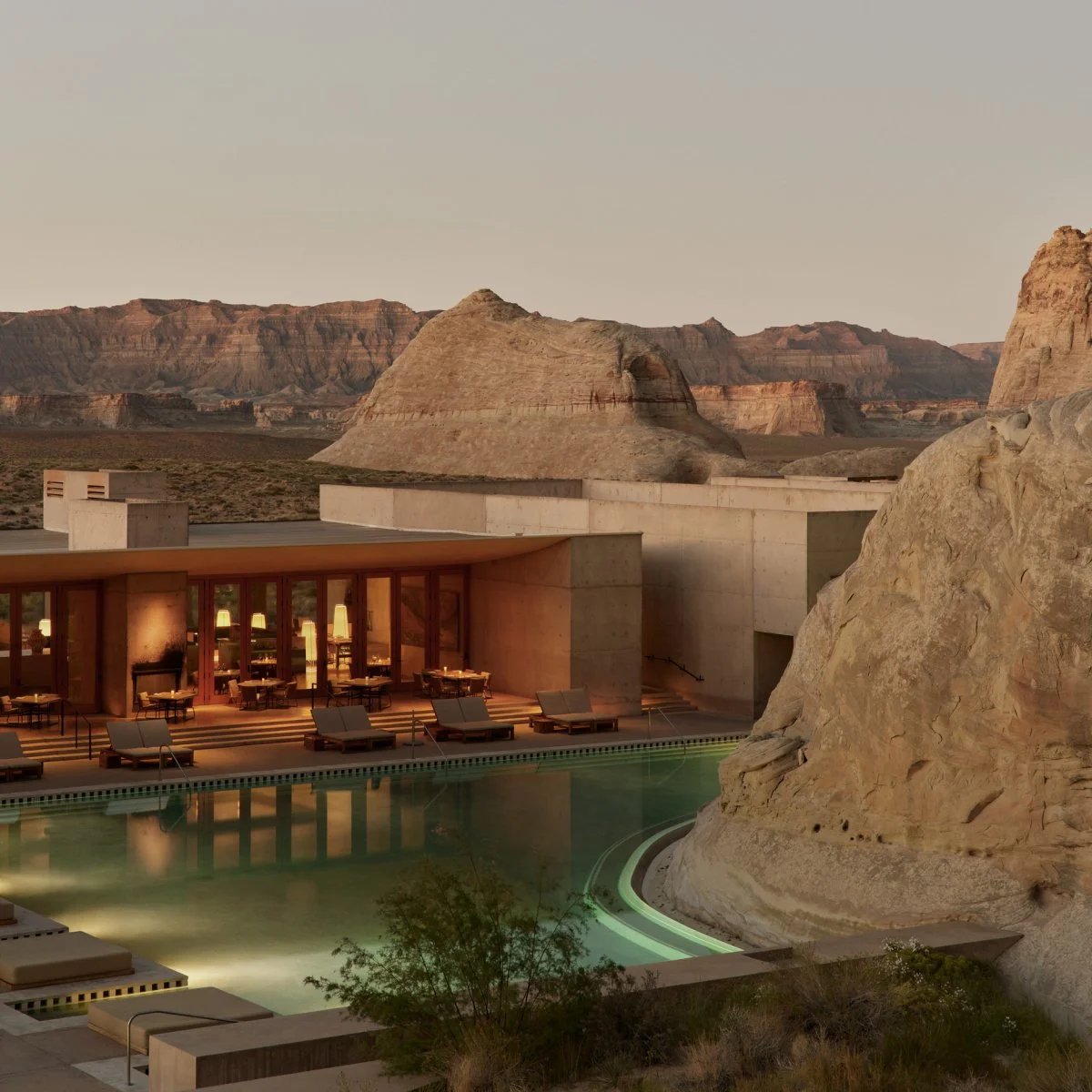 A modern resort with large windows and a lit swimming pool sits among rocky desert formations, with rugged cliffs and clear skies in the background.