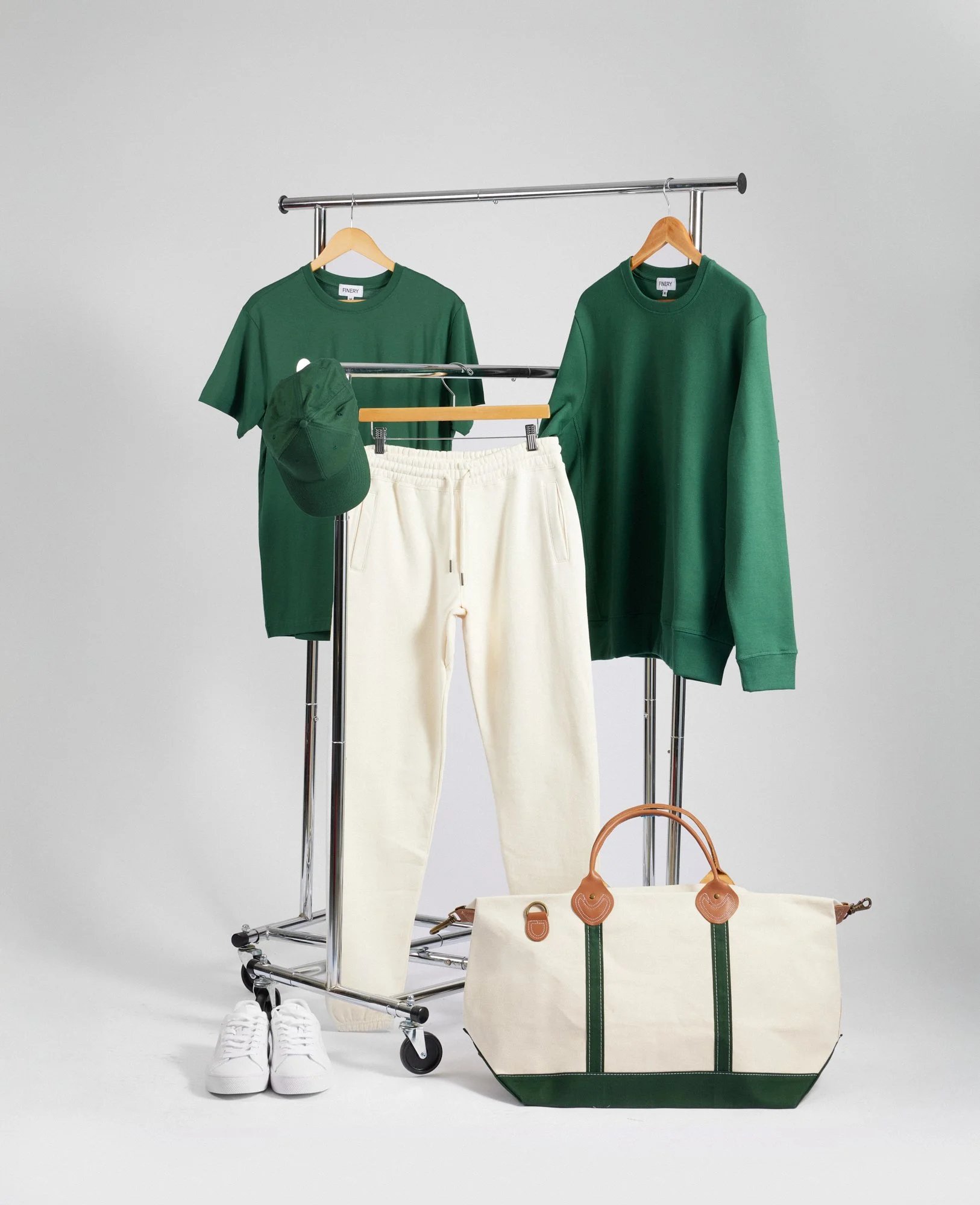 A clothing rack displays a green T-shirt, green sweatshirt, and green cap with cream joggers. White sneakers and a cream tote bag with green accents sit below, all against a plain background.