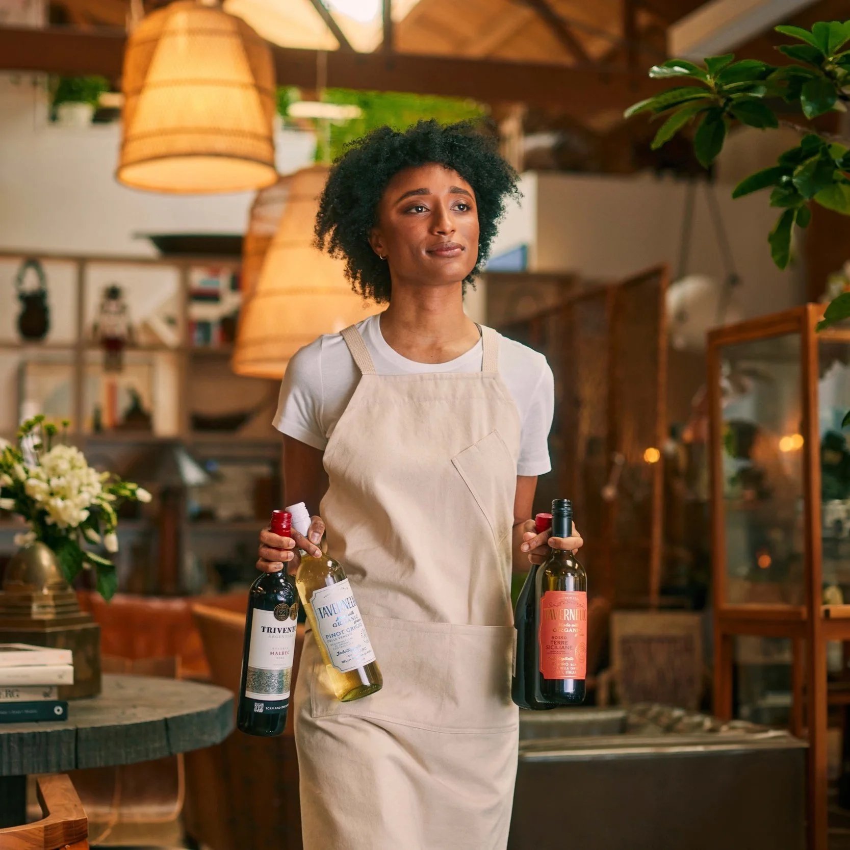 A woman wearing a beige apron and white t-shirt stands indoors, holding three bottles of wine. Warm lighting, plants, and wooden decor give the space a cozy, inviting atmosphere.